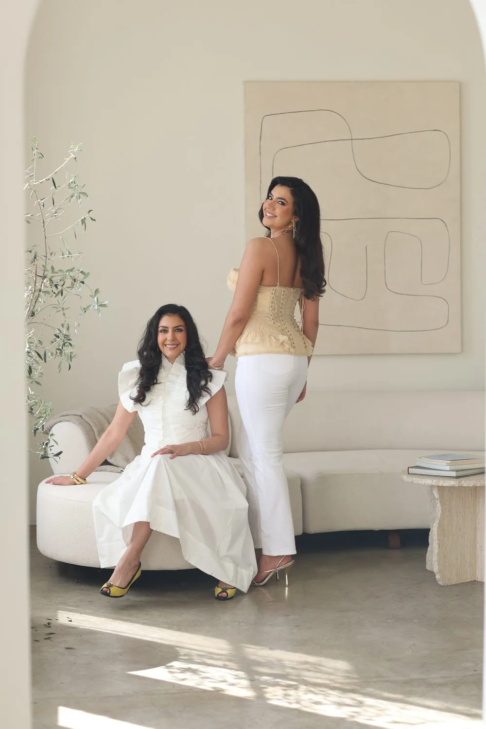 Aurora standing in a cream top and white jeans beside her mother, Dr. Azadeh Khatibi, who sits on a sofa in a white dress.